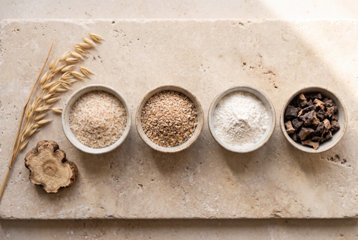 Cuatro cuencos con distintas fuentes de fibra: cáscara de psyllium, salvado de avena, un polvo blanco y trozos de raíz de konjac, junto a una espiga de avena